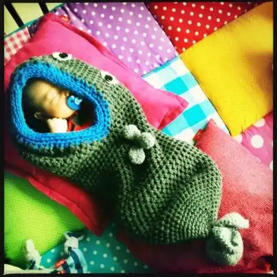 Een slapende baby in een groene gehaakte slaapzak in de vorm van een dier met een blauwe rand.