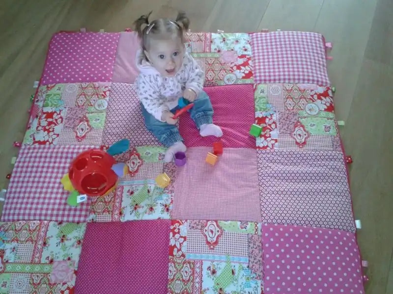Een lachend babymeisje met vlechtjes dat speelt op een kleurrijk patchwork-deken.