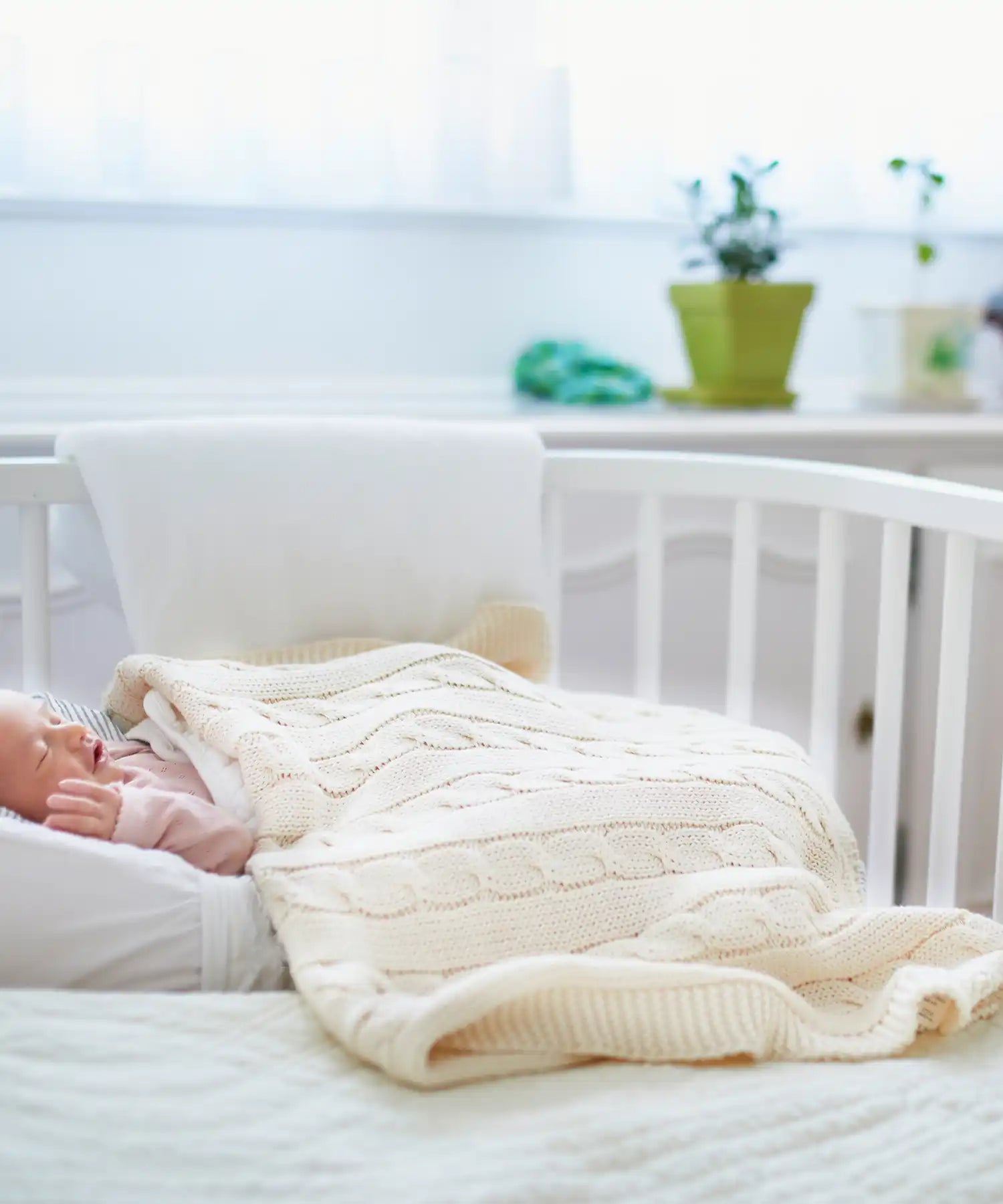 Een baby die slaapt in een witte wieg, bedekt met een roomkleurige kabeltrui deken.