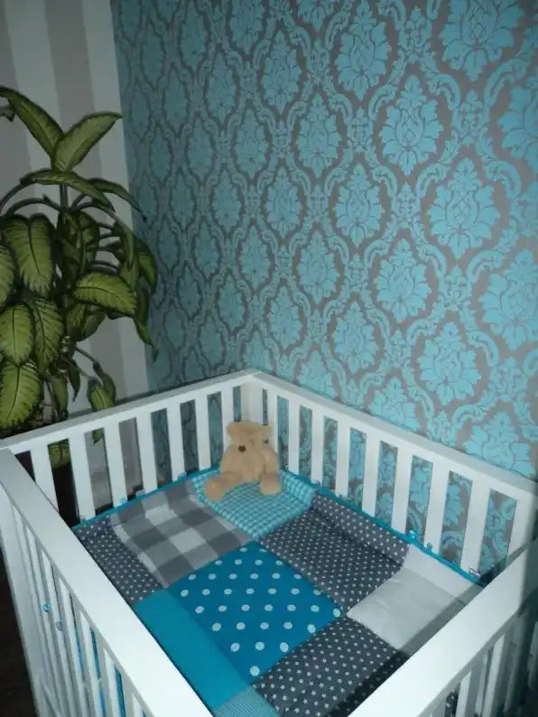 Een witte houten ledikant met een blauw, grijs en wit patchworkdeken en een klein teddybeer.