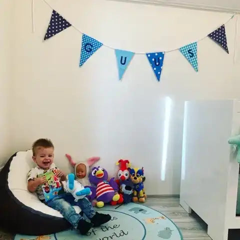 Een jonge jongen zit blij op een grijze en witte zitzak, omringd door een verzameling kleurrijke plush speelgoed.