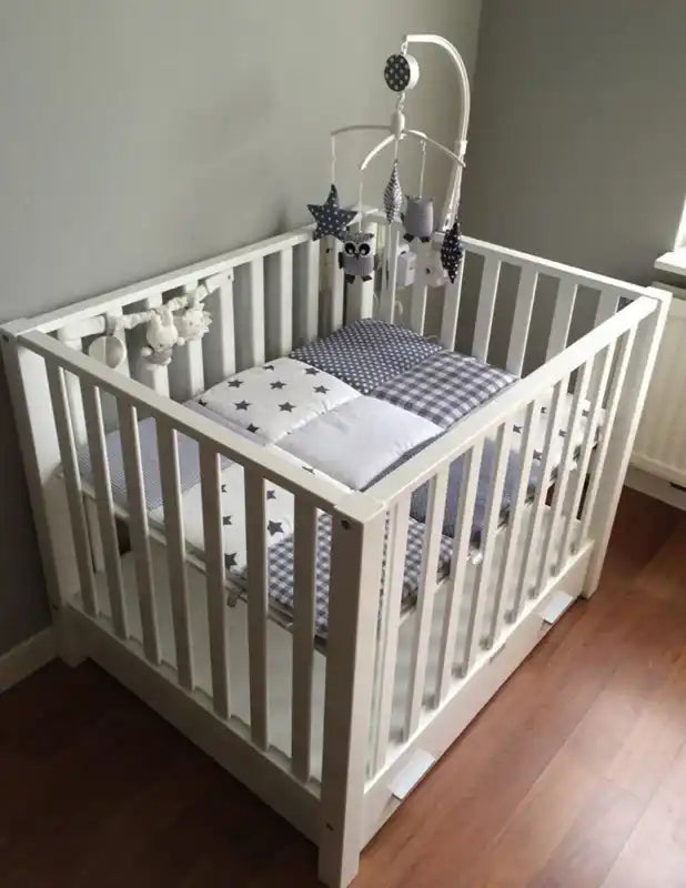 Een witte houten babybedje met grijs en wit beddengoed en een mobiel met ster- en uilversieringen.